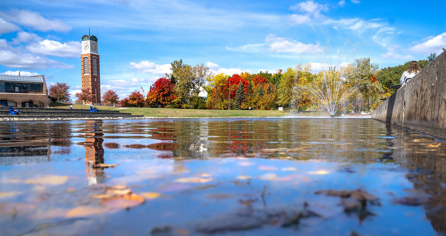 A clock tower stands behind a vibrant autumn tree line, reflected in a shallow pond with a fountain spraying water nearby.